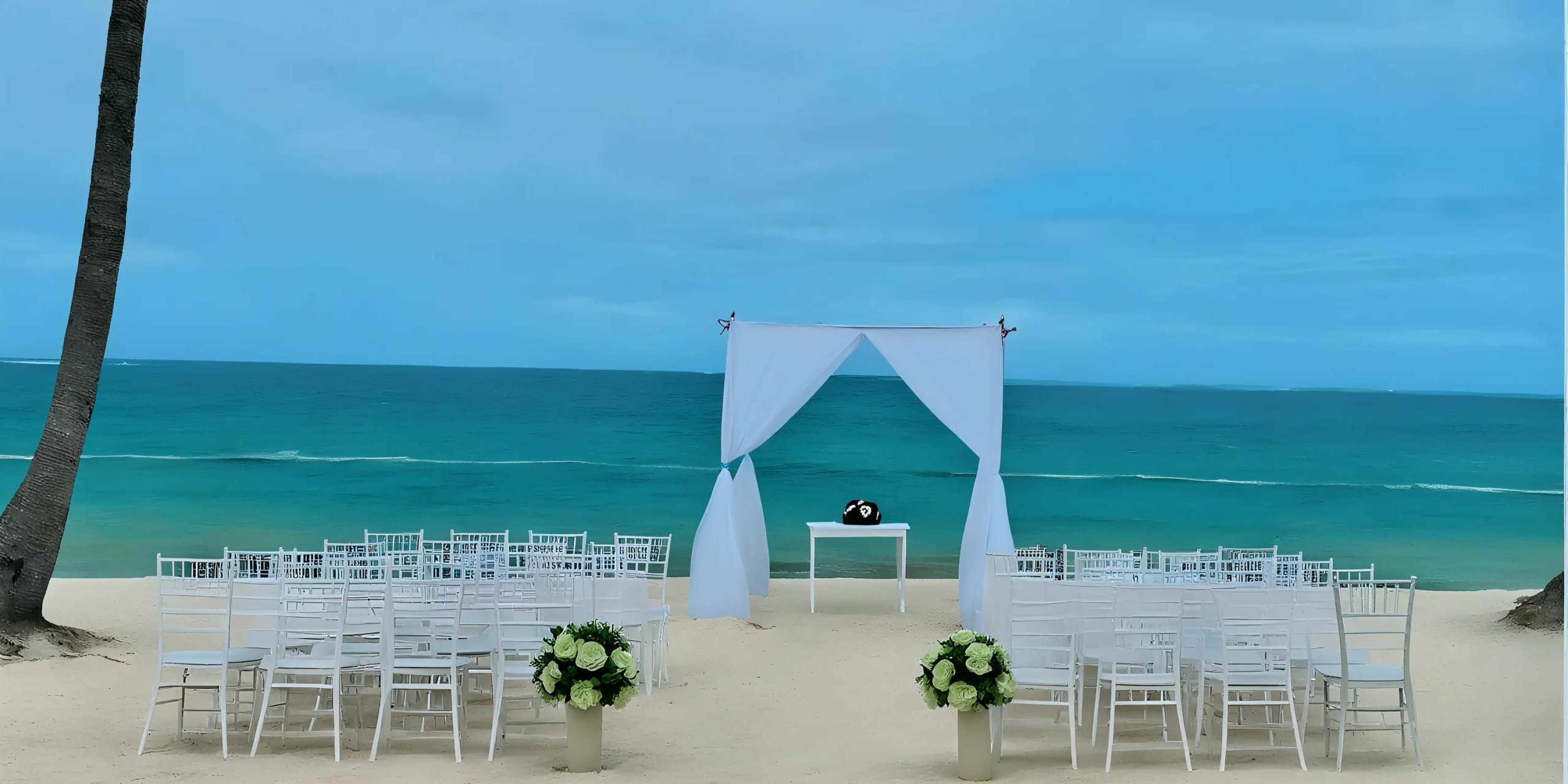 Beach wedding setup with white chairs arranged in rows, a white arch draped with fabric, and a small table with a vase, set against a backdrop of blue ocean and sky.