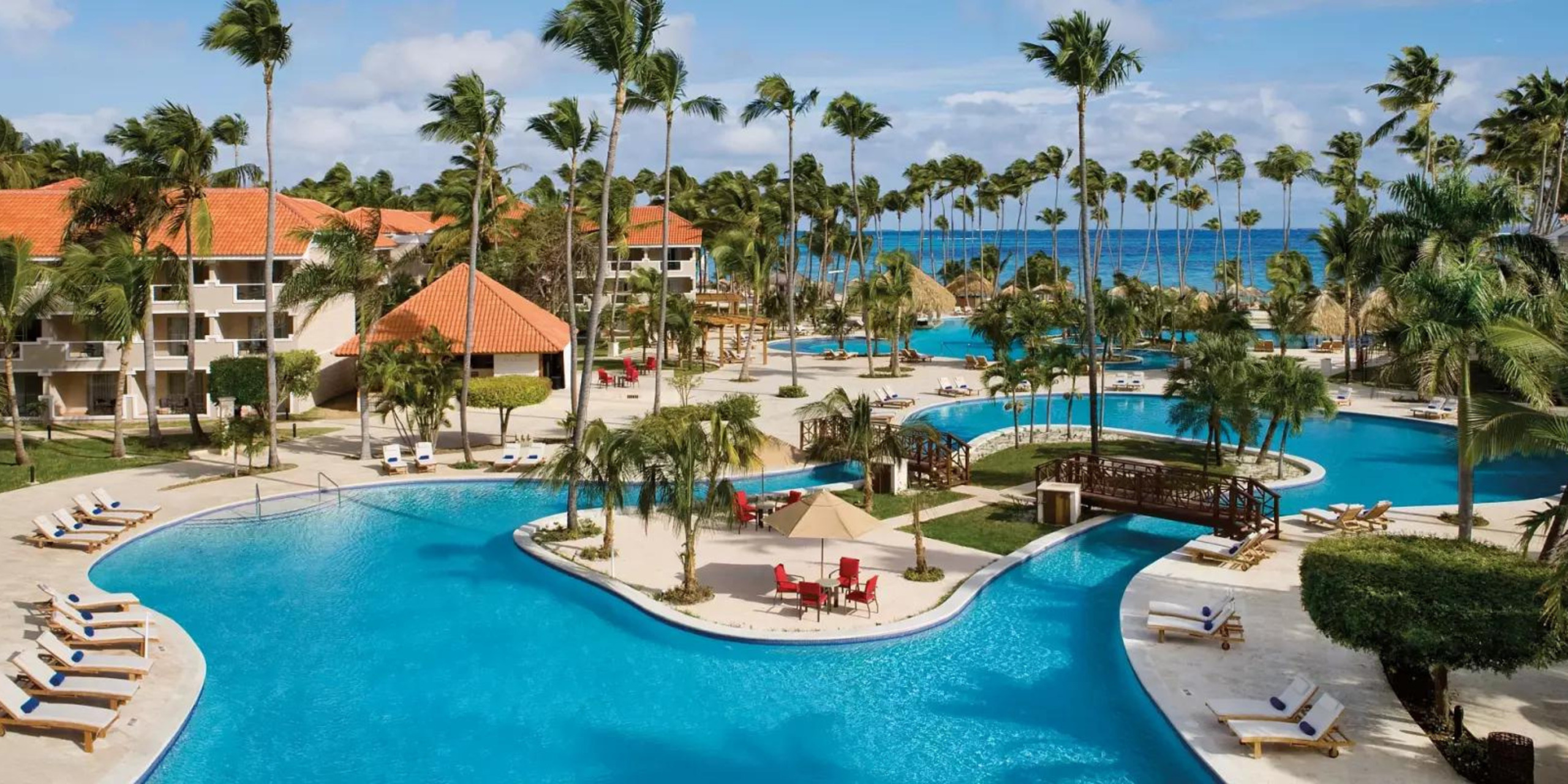 Aerial view of a tropical resort featuring a large curving pool surrounded by palm trees and white buildings, with the Jewel Palm Beach in the background.