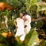 A couple in wedding attire share a moment at their destination wedding, surrounded by lush tropical foliage, viewed through a blurred foreground of leaves.