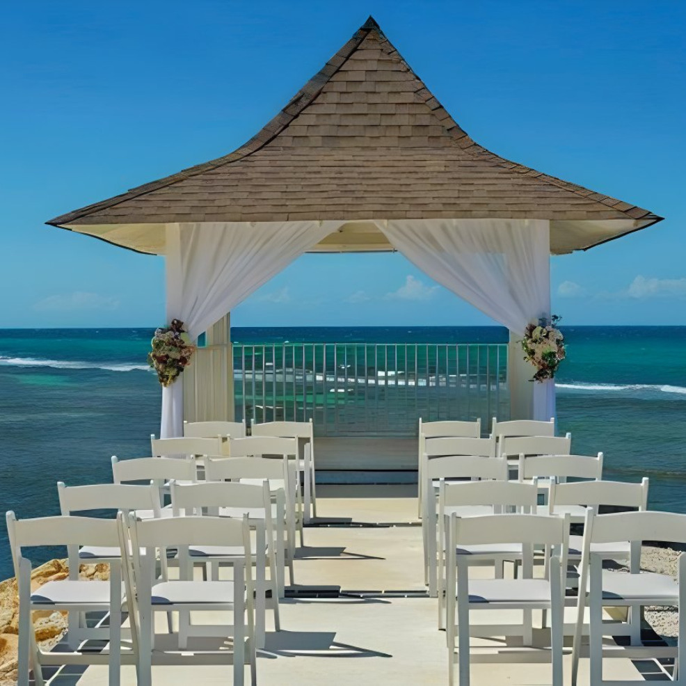 Destination wedding setup with white chairs facing an open gazebo, adorned with floral decorations, overlooking the ocean under a clear blue sky.