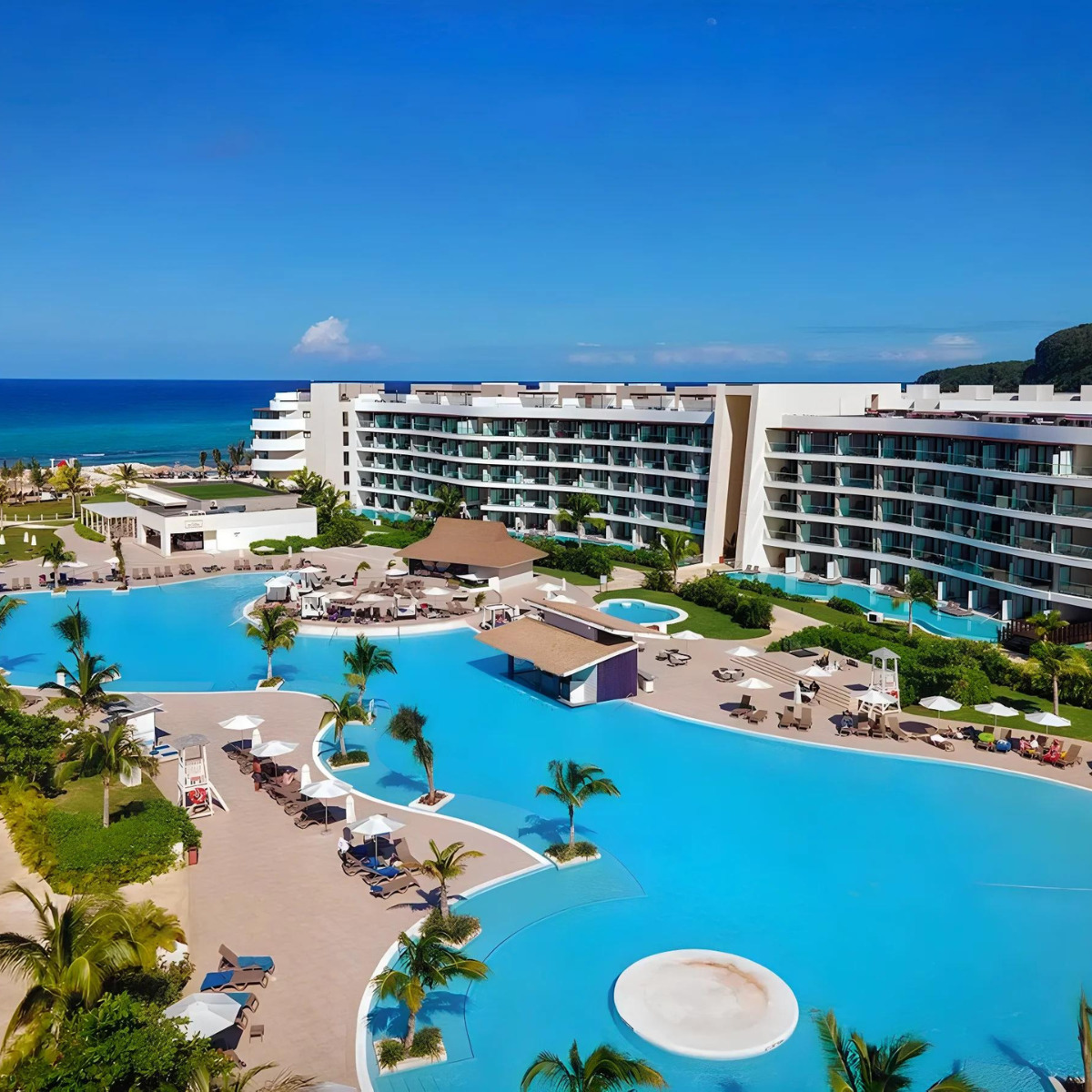 Aerial view of Ocean Coral Spring, a luxury coastal resort, a prime destination for weddings, with expansive swimming pools and modern buildings surrounded by lush greenery and a clear blue sky.