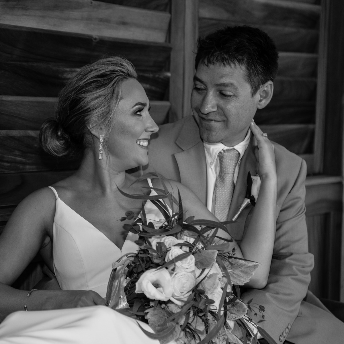 Black and white photo of a smiling bride and groom at their destination wedding, seated closely with the bride holding a bouquet and the groom looking affectionately at her.