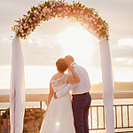A bride and groom stand under a floral arch at their destination wedding, overlooking a sunset at a seaside, in an intimate pose with the groom kissing the bride’s head.