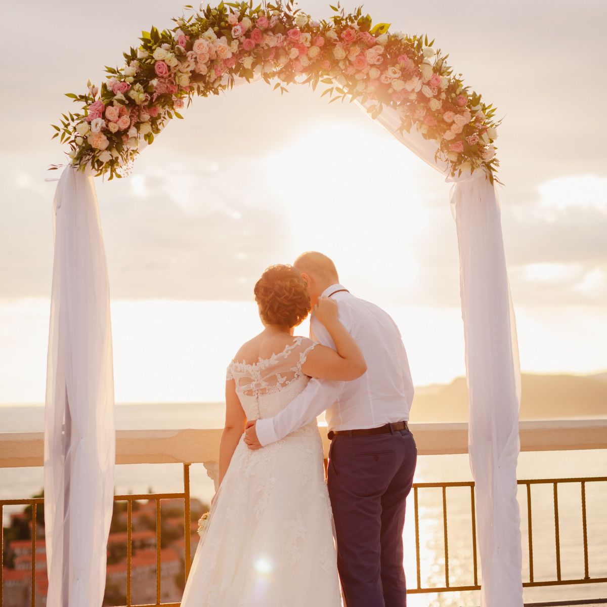 A bride and groom stand under a floral arch at their destination wedding, overlooking a sunset at a seaside, in an intimate pose with the groom kissing the bride’s head.