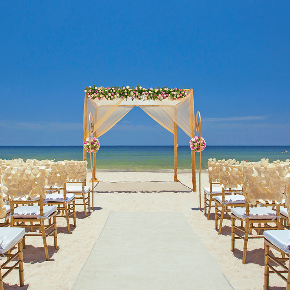 Destination wedding setup with a flower-adorned arch, white chairs, and a central aisle leading to the ocean under a clear blue sky.