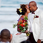 A couple in wedding attire sharing a kiss on a beach at their destination wedding; the bride holds a colorful bouquet, and an onlooker is partially visible in the foreground.