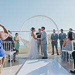 A bride and groom exchanging vows at a destination wedding ceremony, surrounded by guests under a clear blue sky.