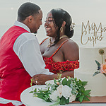 A joyful couple at their destination wedding, in wedding attire closely embracing beside a cake with "mr & mrs curtis" written on it, against a sunset ocean backdrop.
