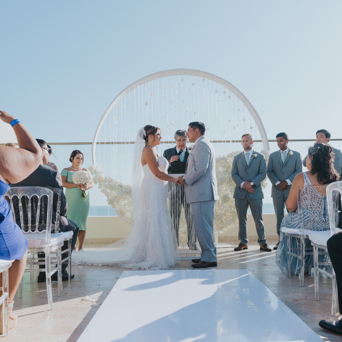 A bride and groom exchanging vows at a destination wedding ceremony, surrounded by guests under a clear blue sky.