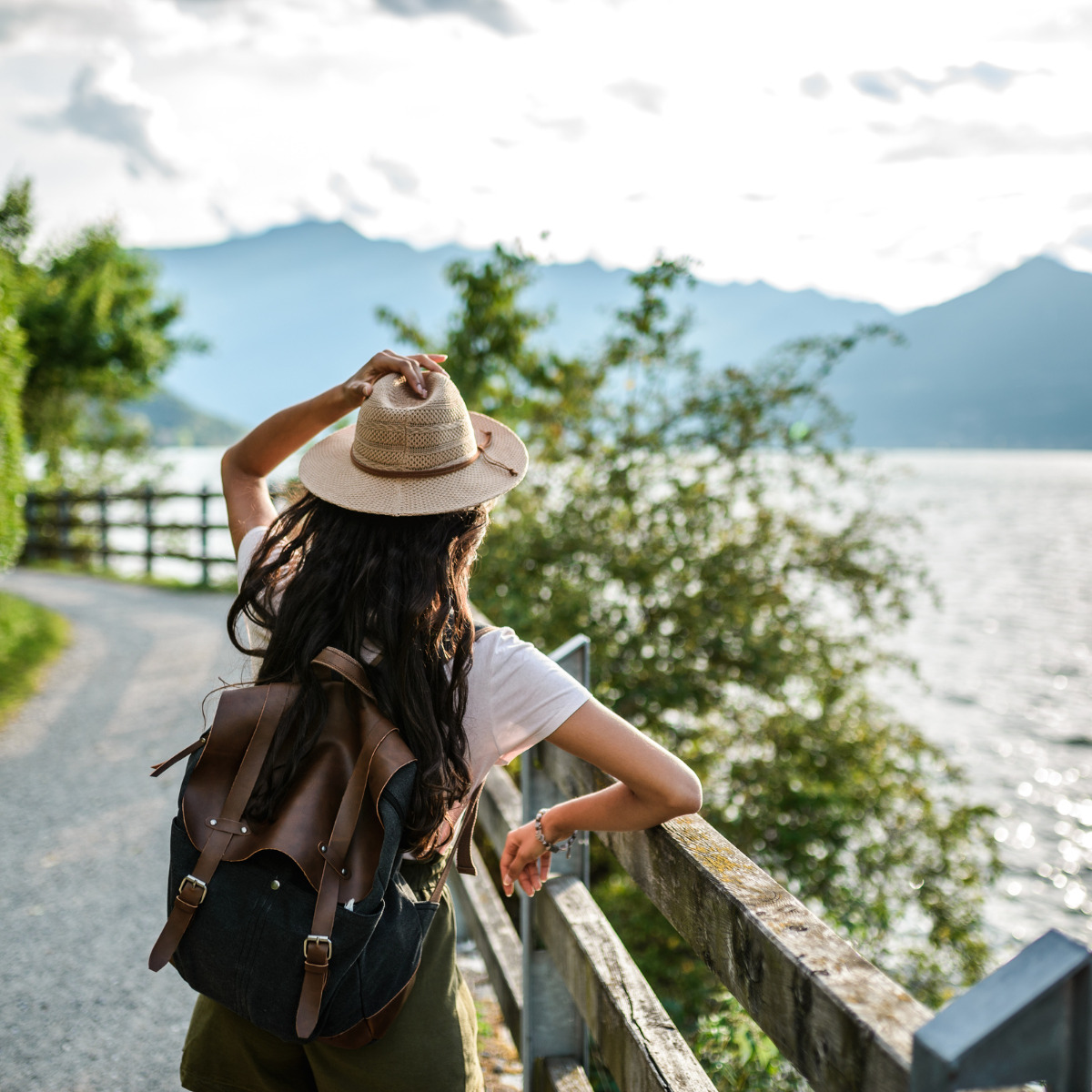 Woman with a backpack and straw hat looking over a lake, standing next to a wooden fence on a path with mountain views in the background, scouting for her destination wedding location.