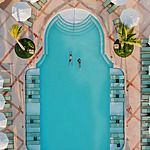 Aerial view of two people swimming in a turquoise pool shaped like a keyhole, surrounded by white sun loungers and green plants at a destination wedding venue.