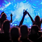 Silhouettes of people at a concert raising their hands, with a blue stage light in the background.