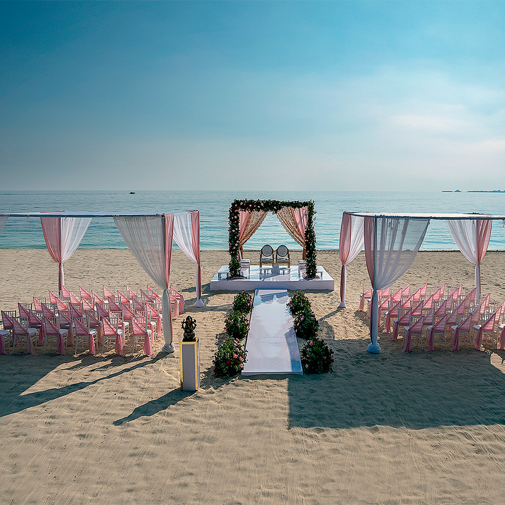 Beach wedding setup with a white altar and pink drapery. Rows of chairs with pink covers are arranged on both sides of an aisle leading to the ocean view.