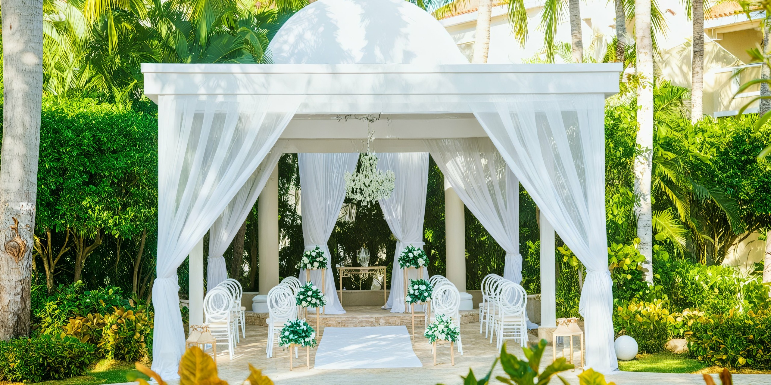 Experience the perfect Jewel Palm Beach wedding at an outdoor venue featuring a charming white gazebo adorned with sheer curtains, enveloped by lush greenery. Elegant white chairs and stunning floral arrangements line the aisle, culminating in a breathtaking chandelier inside the gazebo—making your special day unforgettable.