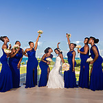 A bride in a white dress with six bridesmaids in blue dresses, laughing and posing together outdoors by the sea at a Destify wedding.