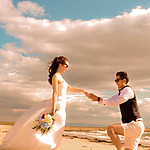 A bride in a white dress and a groom in a vest holding hands on a beach under a cloudy sky, with the groom kneeling and the bride twirling, perfectly capturing the essence of destination weddings.