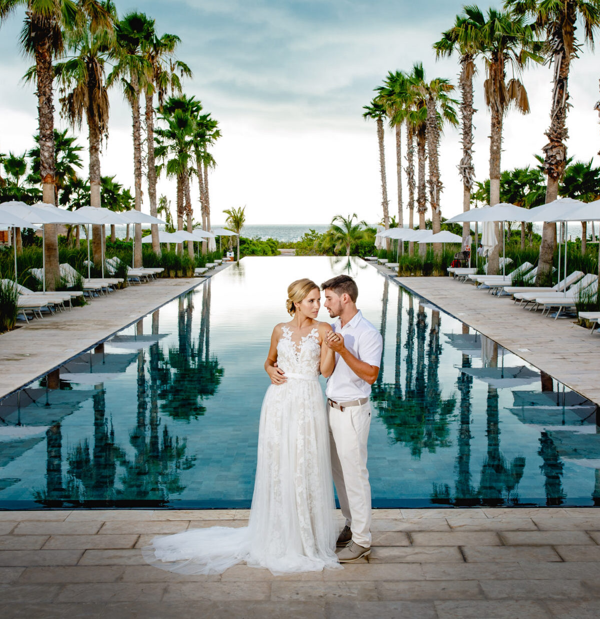 A bride and groom posing in front of a pool with palm trees for their destination wedding.