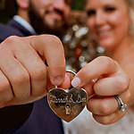 A couple at their destination wedding holding a heart-shaped lock engraved with "hannah & ben," with a blurry background featuring their smiling faces.