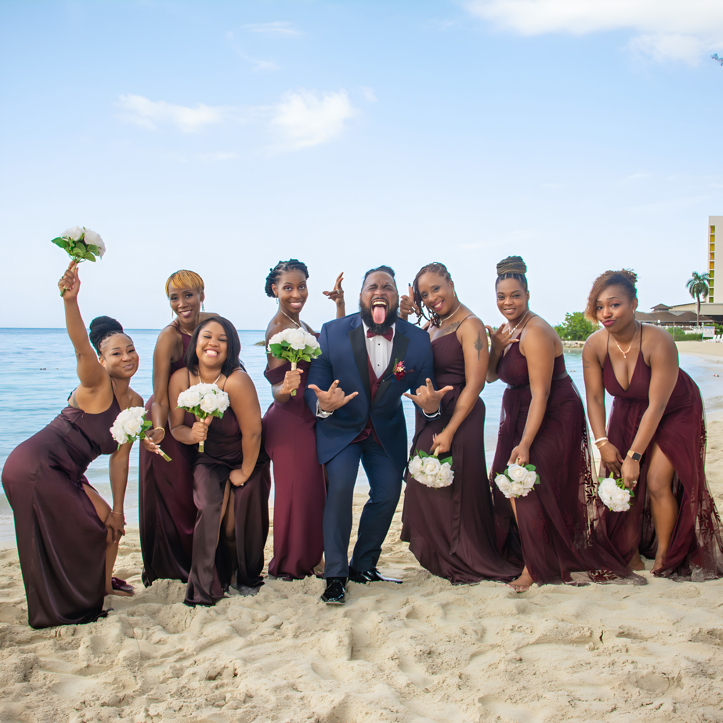 A groom in a blue suit joyfully poses on a beach during a destination wedding with seven bridesmaids in matching maroon dresses, all holding bouquets.