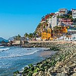 A scenic coastal view featuring a rocky shore, blue sea, and a clifftop lined with colorful buildings under a clear sky, perfect for destination weddings.
