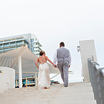 A bride and groom walking hand in hand up a flight of stairs, with buildings in the background under a cloudy sky.