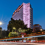 Modern high-rise building illuminated with purple lights at dusk, a popular venue for destination weddings, with light trails from passing traffic on the street in front.