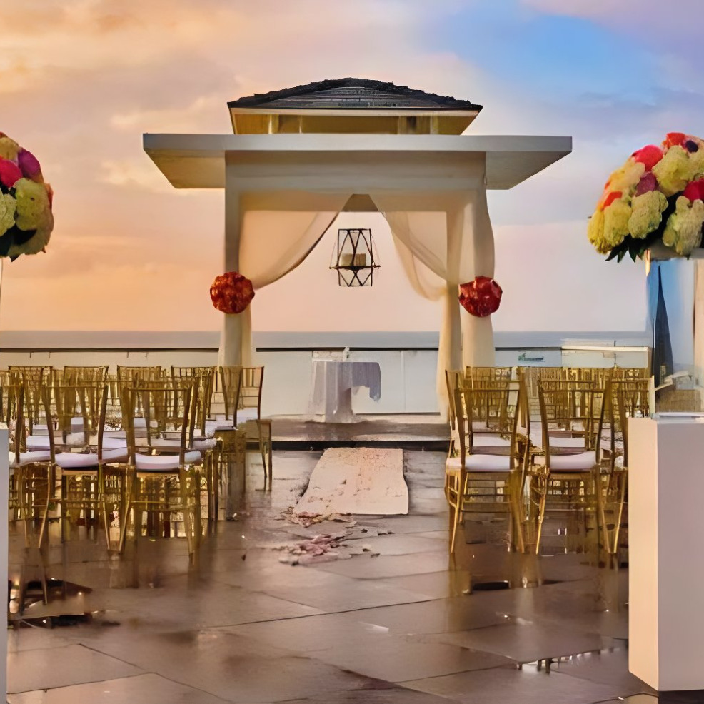 Destination wedding setup at sunset with a white gazebo, chairs lined up, and floral decorations, overlooking the ocean.