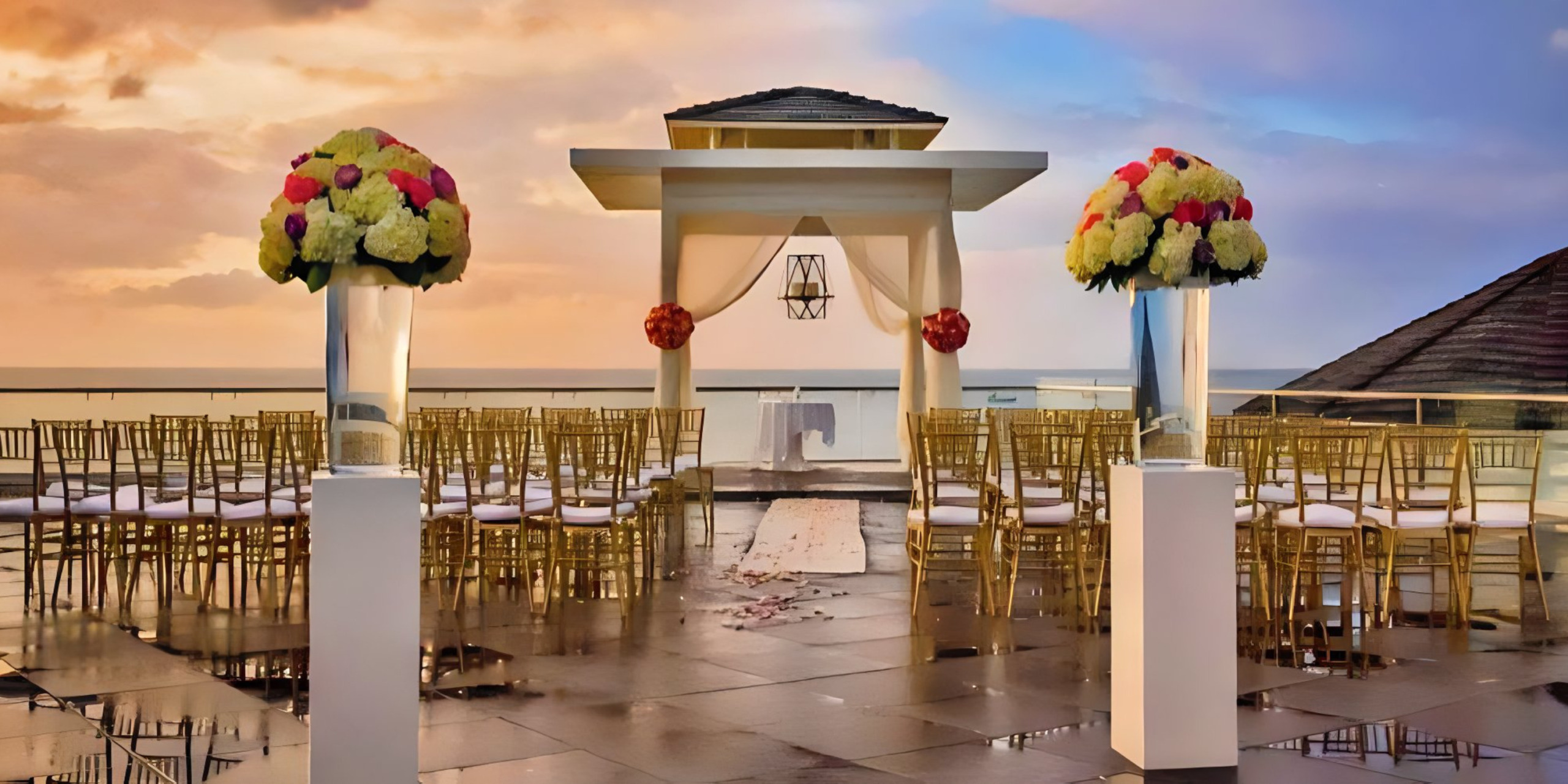 Destination wedding setup at sunset with a gazebo, flower-adorned pedestals, and rows of gold chairs facing the ocean.