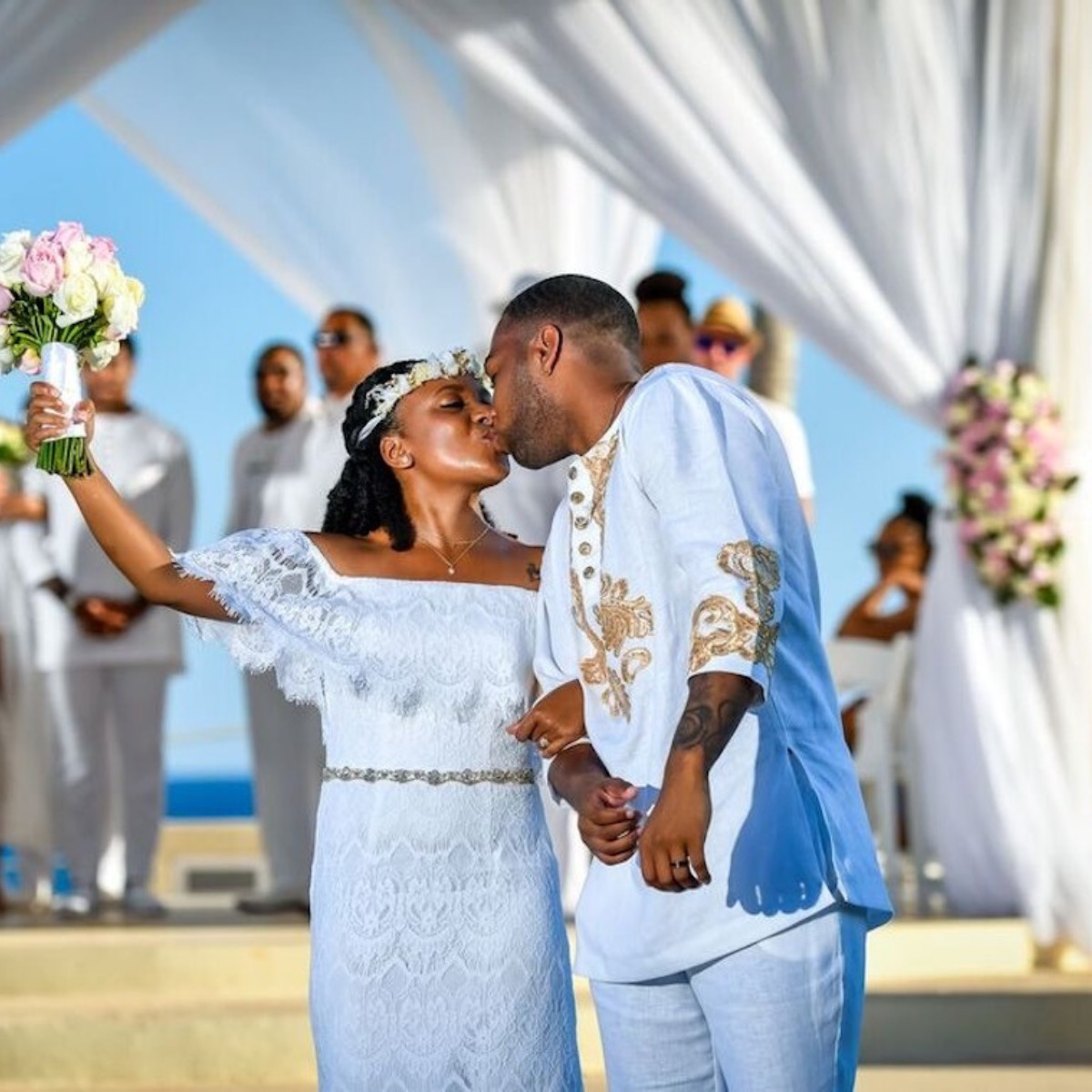 A bride and groom kissing at their destination wedding ceremony, dressed in white and blue, under a white canopy with guests in the background.