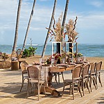 Elegant outdoor dining setup on a wooden deck by the sea, perfect for destination weddings, featuring a table with floral centerpieces, surrounded by wicker chairs under tall palm trees.