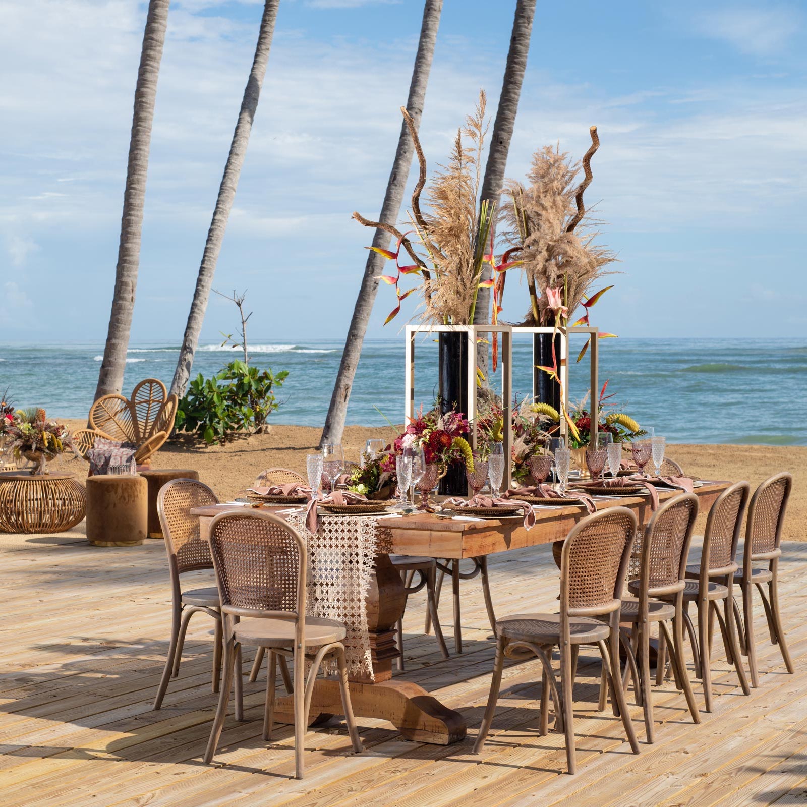 Elegant outdoor dining setup on a wooden deck by the sea, perfect for destination weddings, featuring a table with floral centerpieces, surrounded by wicker chairs under tall palm trees.