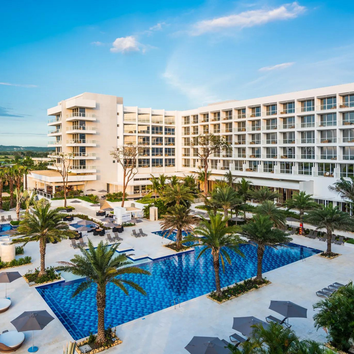 A modern resort with an expansive outdoor swimming pool, perfect for a destination wedding, surrounded by palm trees, loungers, and a spacious white building under a clear sky.
