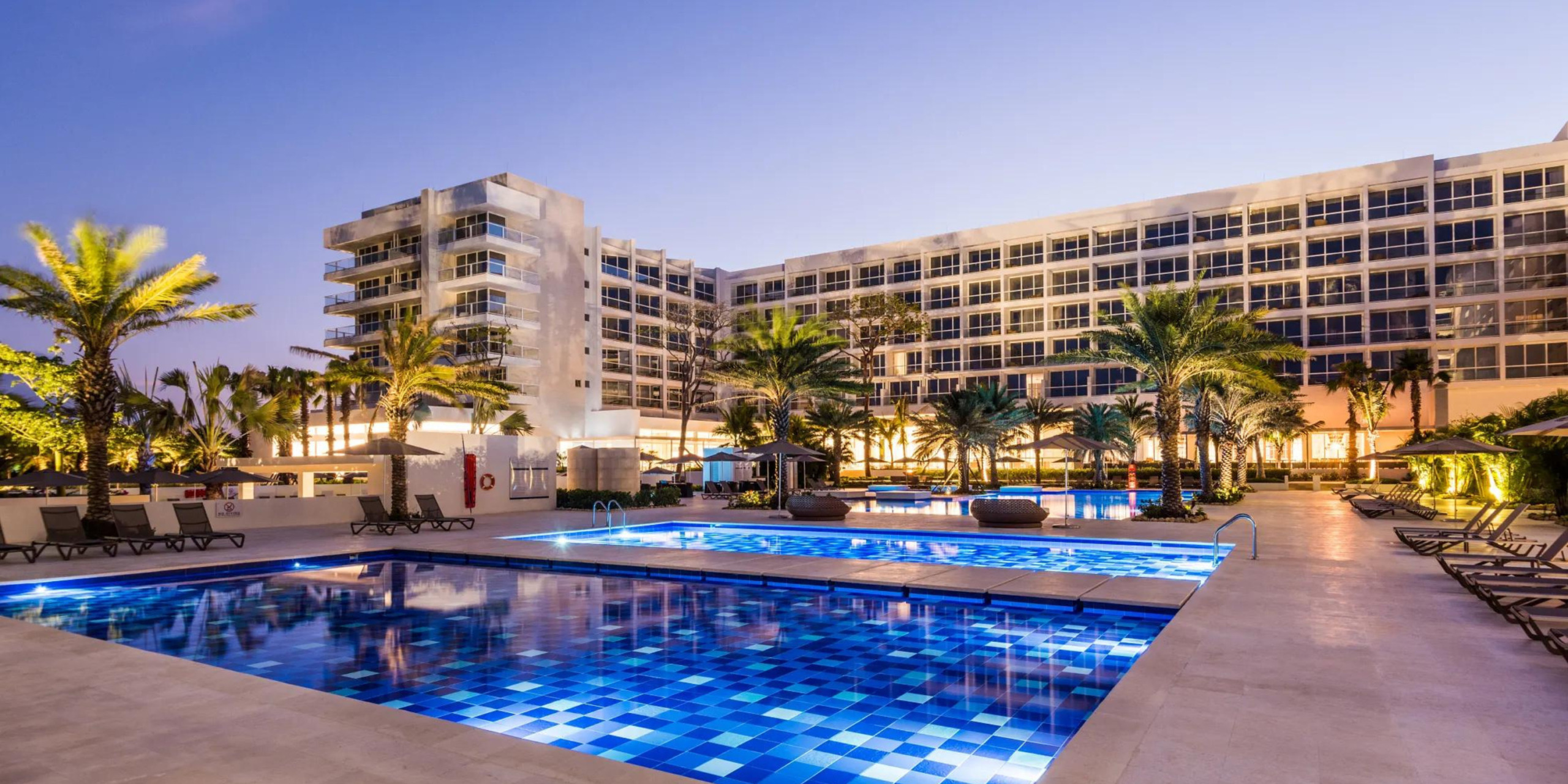 Dreams Karibana Cartagena Golf & Spa Resort exterior at twilight, a perfect destination for weddings, with an illuminated swimming pool surrounded by palm trees and modern building façade.