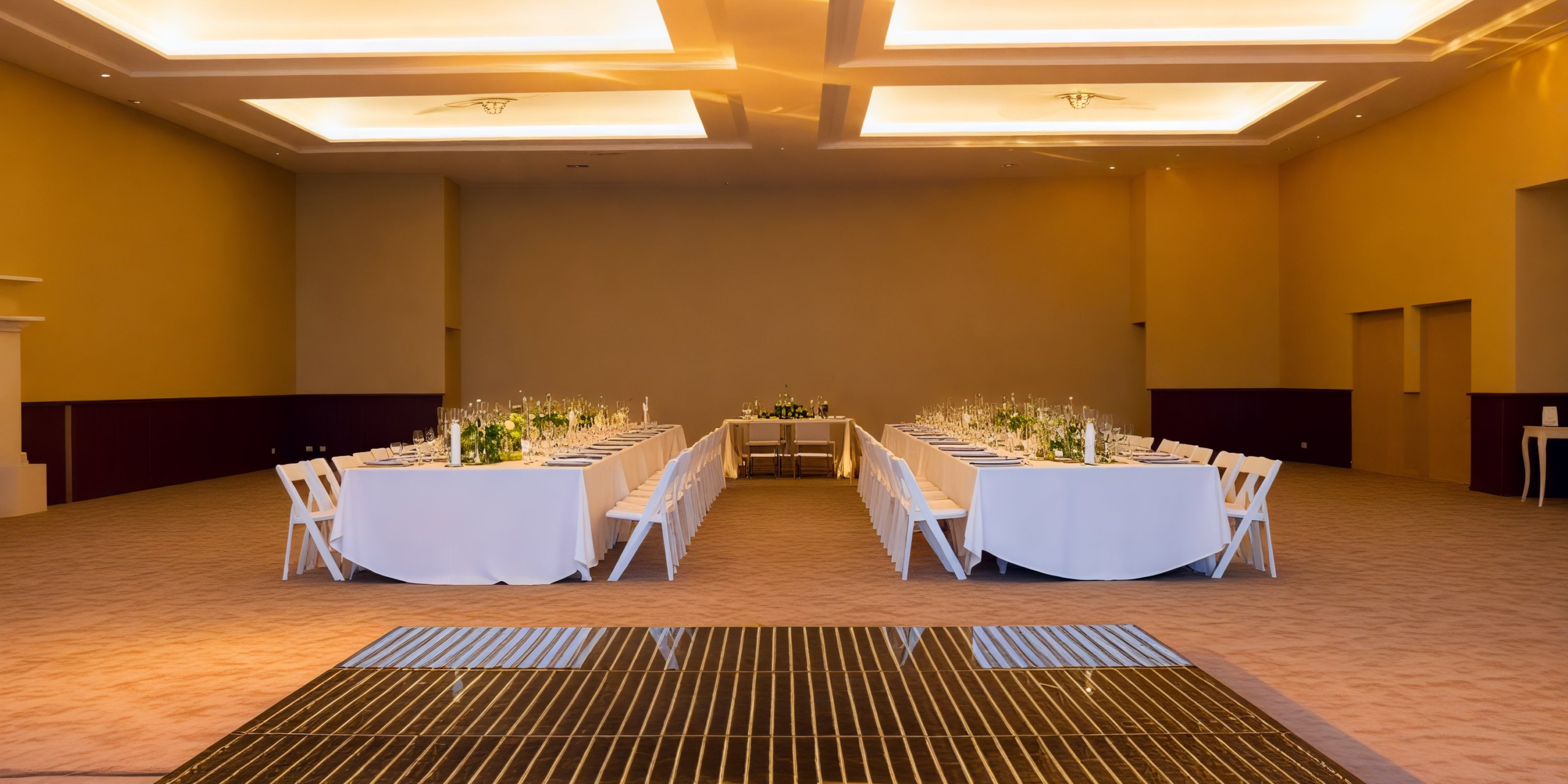 A spacious banquet hall is elegantly arranged for a destination wedding, featuring two long tables draped with pristine white tablecloths.