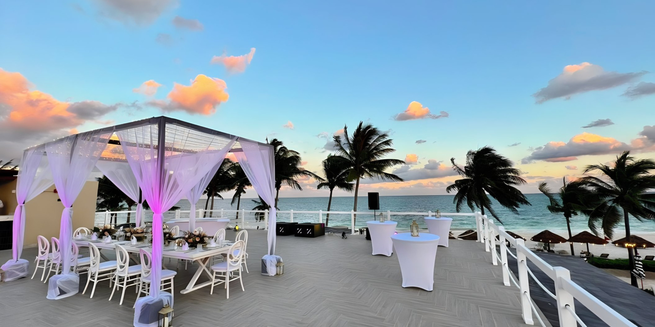 Outdoor destination wedding setup on a terrace overlooking the ocean at sunset, featuring white chairs and draped canopy, with palm trees and a cloudy sky in the background.
