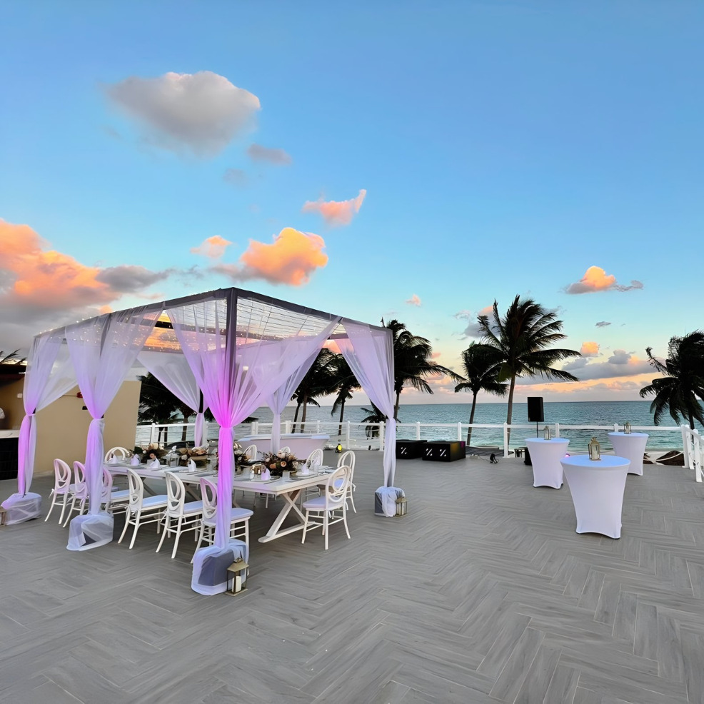 Destination wedding setup on a beach at sunset, featuring a white canopy, decorated tables, and palm trees against a backdrop of colorful clouds.