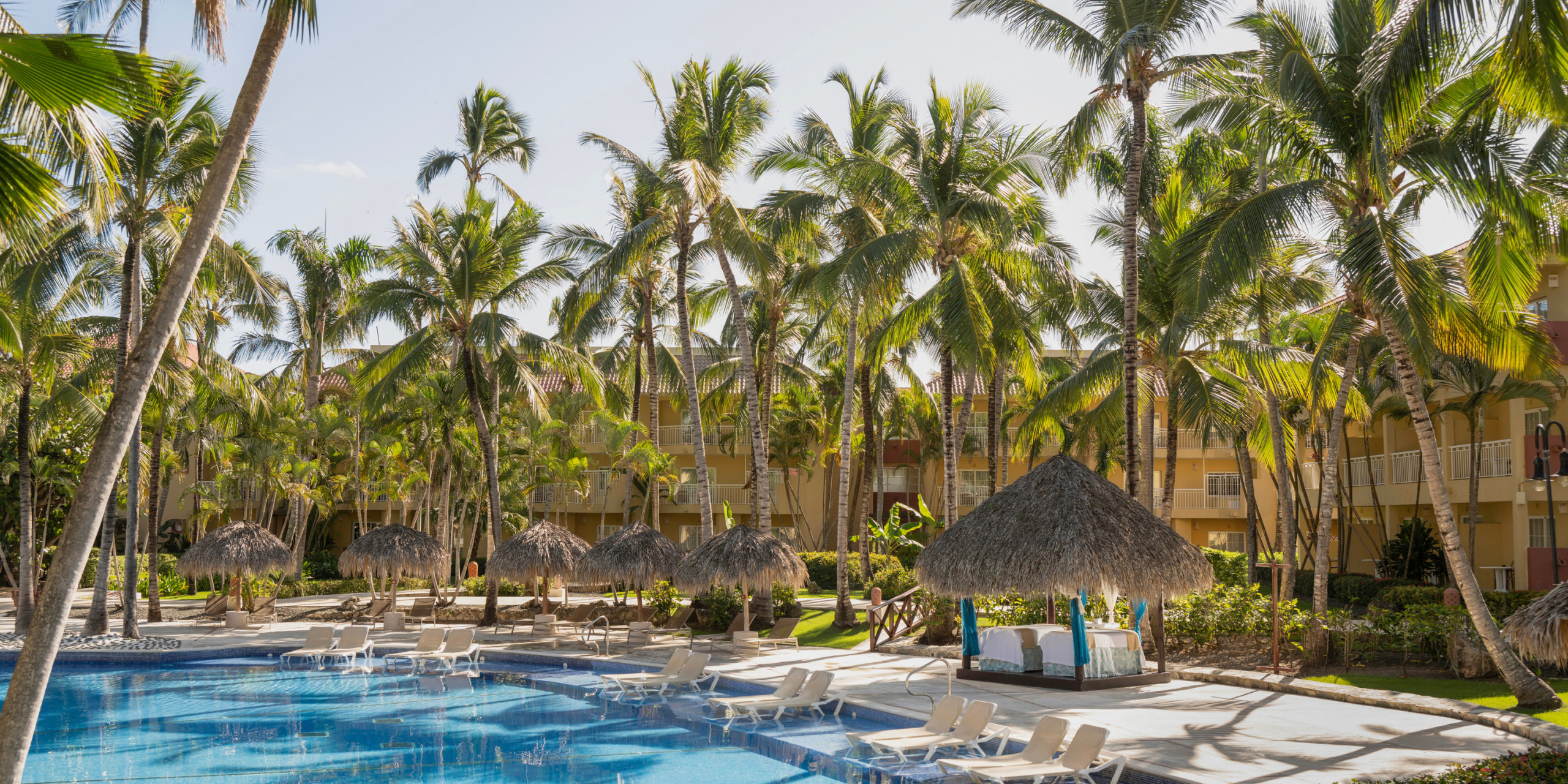 Jewel Punta Cana, a lush tropical resort, a perfect destination for weddings, with a swimming pool surrounded by palm trees and straw umbrellas, adjacent to a multi-story hotel building.