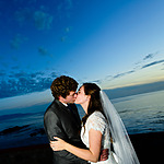 A newlywed couple kissing at sunset by the sea under a blue sky with clouds during their destination wedding.