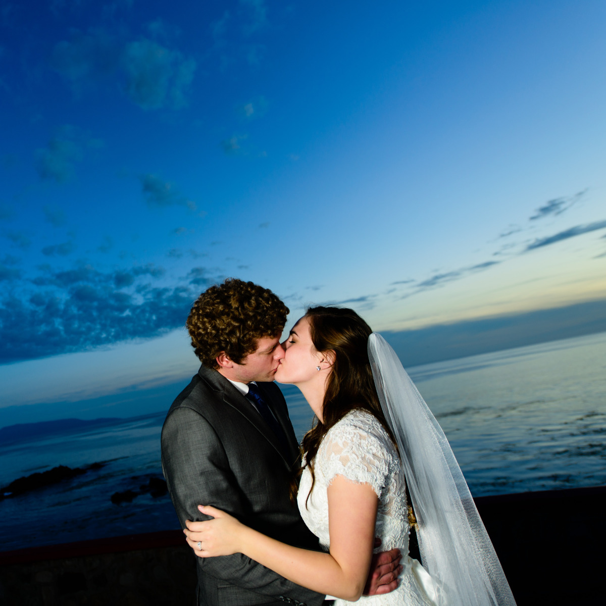 A newlywed couple kissing at sunset by the sea under a blue sky with clouds during their destination wedding.