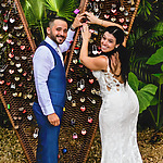 A newlywed couple at a destination wedding joyfully locks a padlock on a heart-shaped installation covered with other locks, symbolizing lasting love.