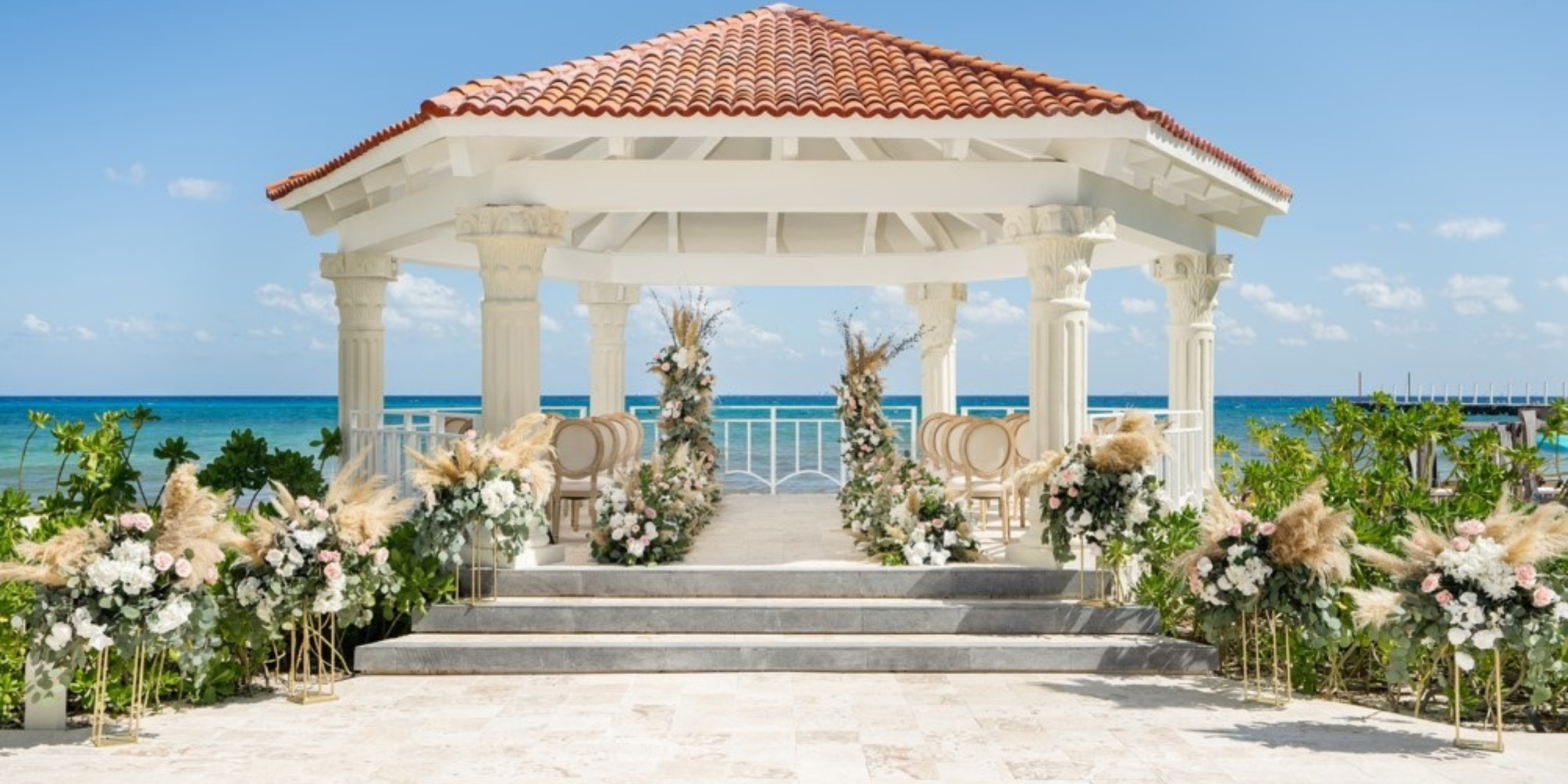 Destination oceanfront gazebo decorated with floral arrangements for a wedding, with clear skies and calm sea in the background.