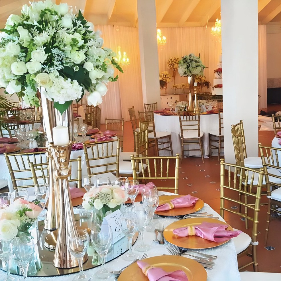 Elegant destination wedding reception setup featuring tall floral centerpiece, gold chairs, and tables adorned with pink napkins and clear glassware.
