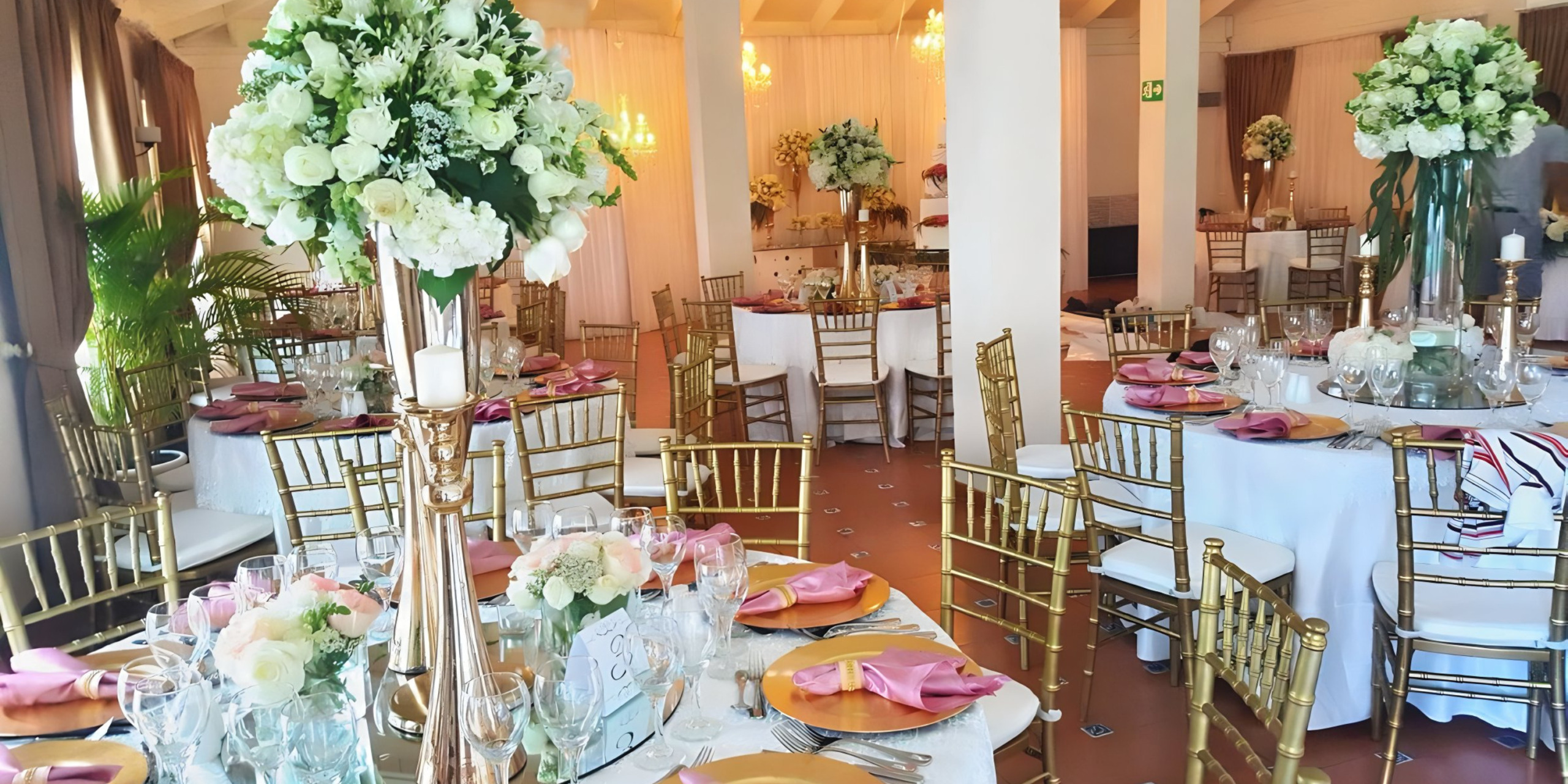 Elegant destination wedding reception hall with floral centerpieces, gold chairs, and tables set with pink napkins and crystal glassware.