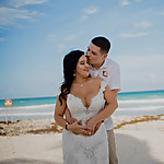 A groom in a white shirt kisses the forehead of a bride in a lace gown on a beach with clear blue skies and turquoise waters, perfect for destination weddings.
