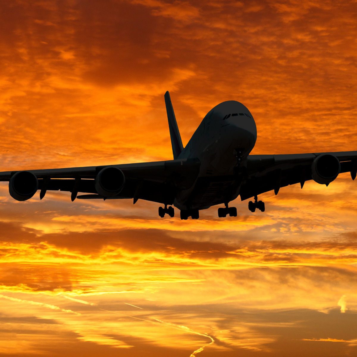 A commercial airplane silhouetted against a vibrant orange sunset sky, descending for landing with extended landing gear, carrying guests to a destination wedding.