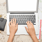 Top-down view of a person's hands typing on a laptop placed on a desk scattered with coins, a wallet, receipts, and a map of destination weddings.