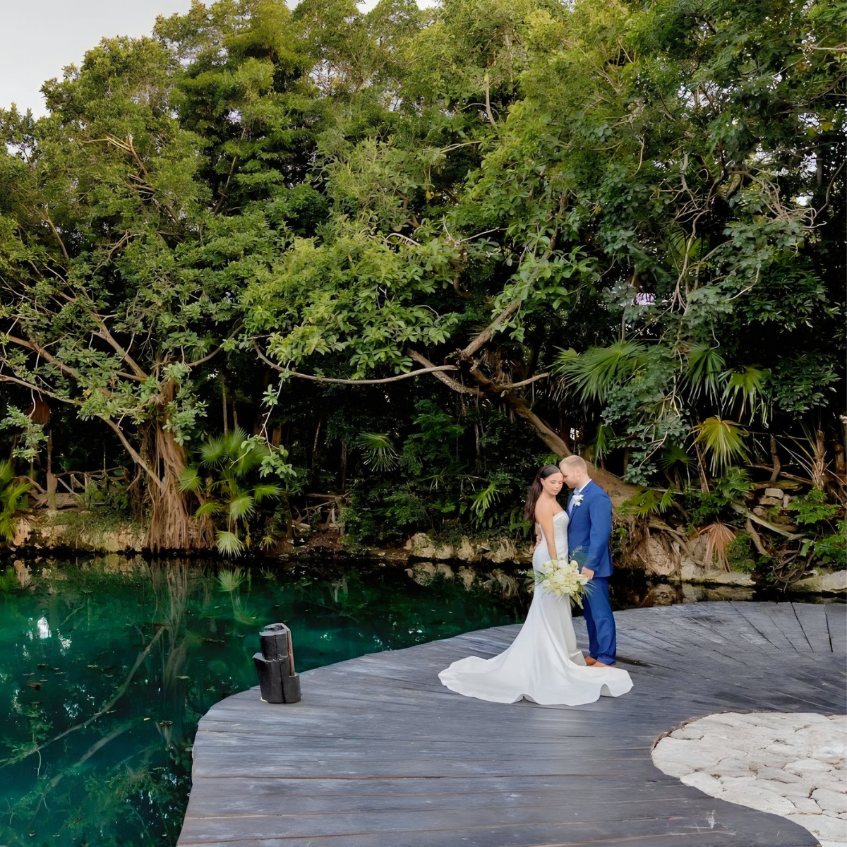 A bride and groom share a kiss on a wooden dock by a serene natural pond surrounded by lush greenery, perfect for destination weddings.