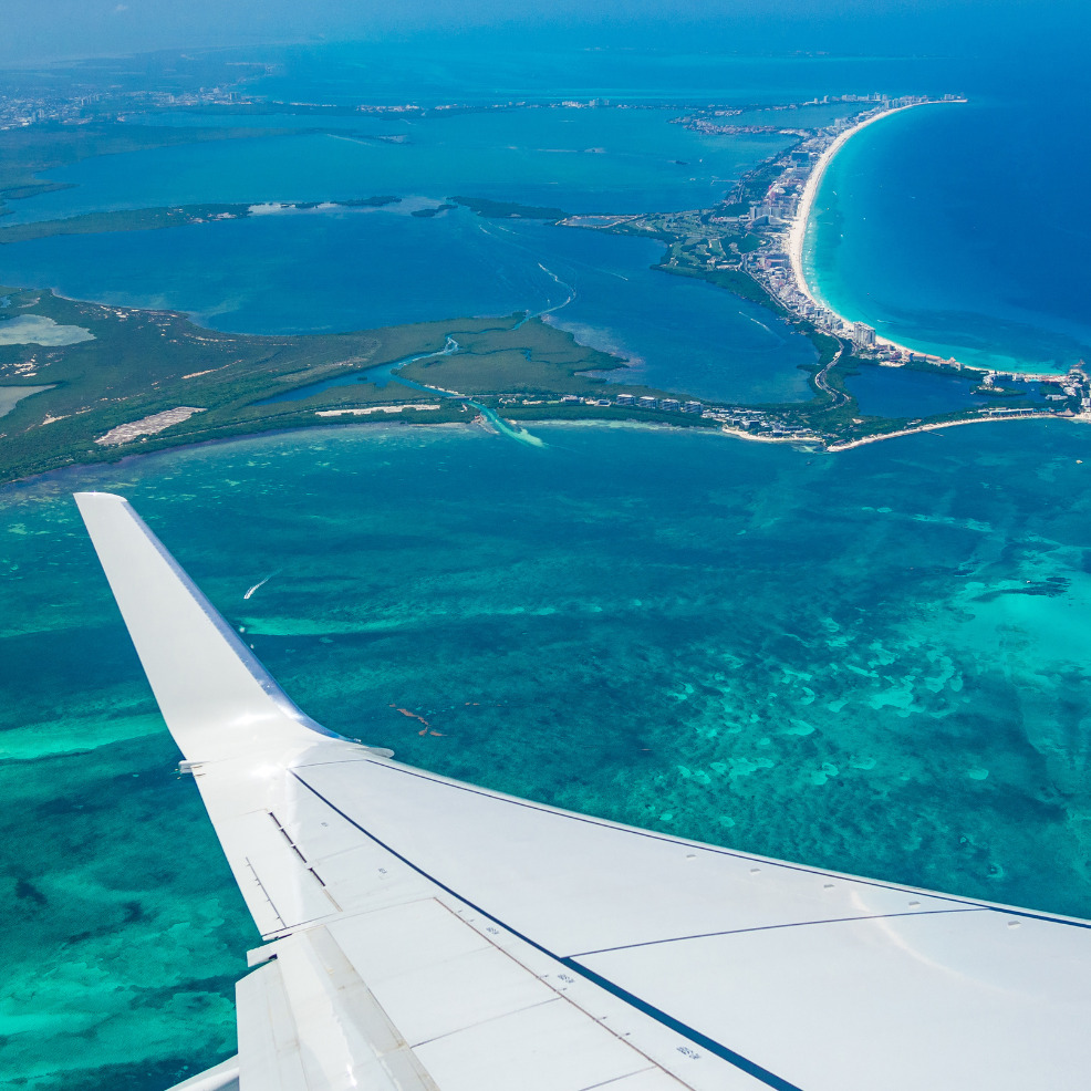 Aerial view from an airplane showing the wing over vibrant turquoise ocean waters near a coastal city, a popular destination wedding locale.