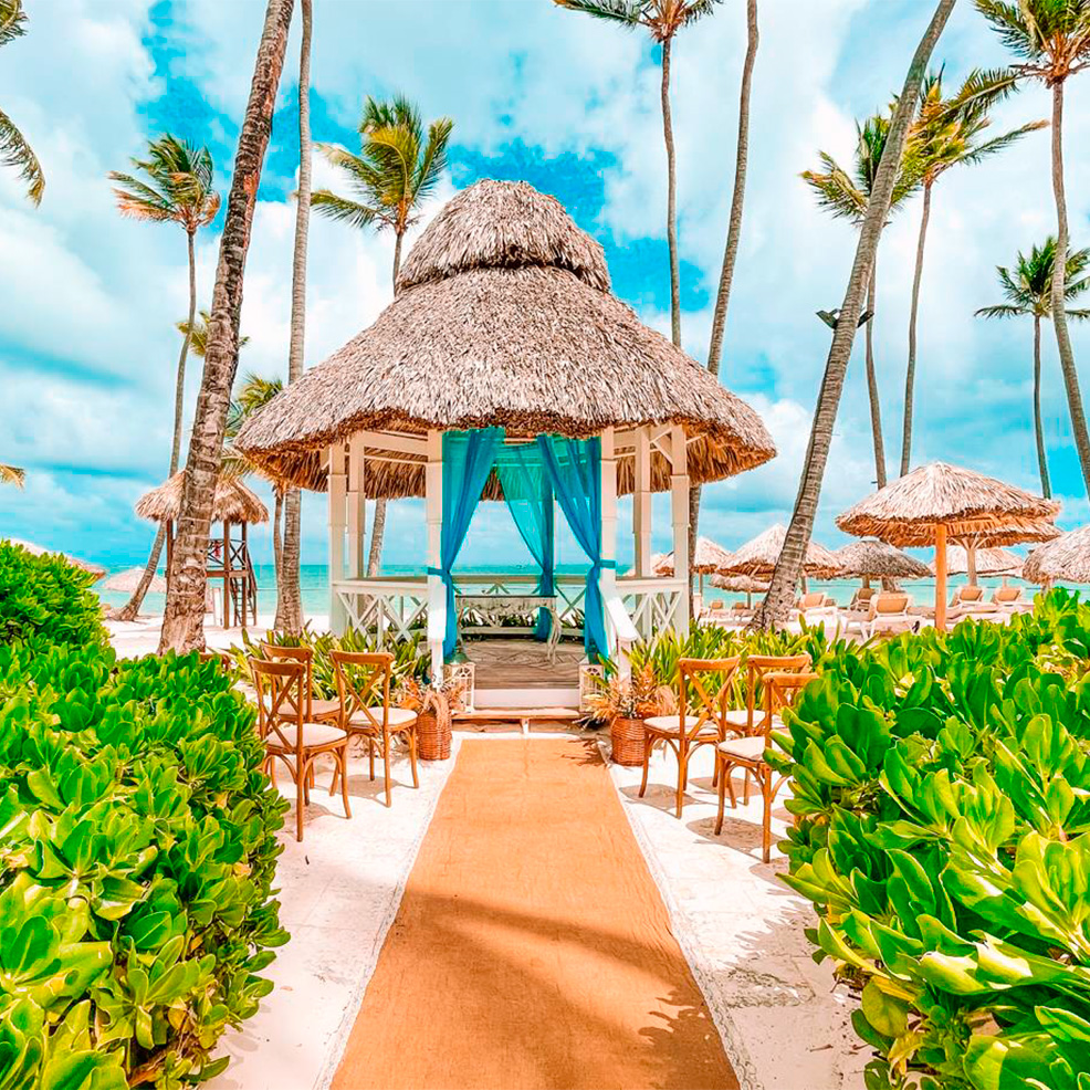 A beachside gazebo with a thatched roof and blue drapes is surrounded by green shrubs. Wooden chairs line a sandy pathway leading to it, with palm trees and the ocean in the background.