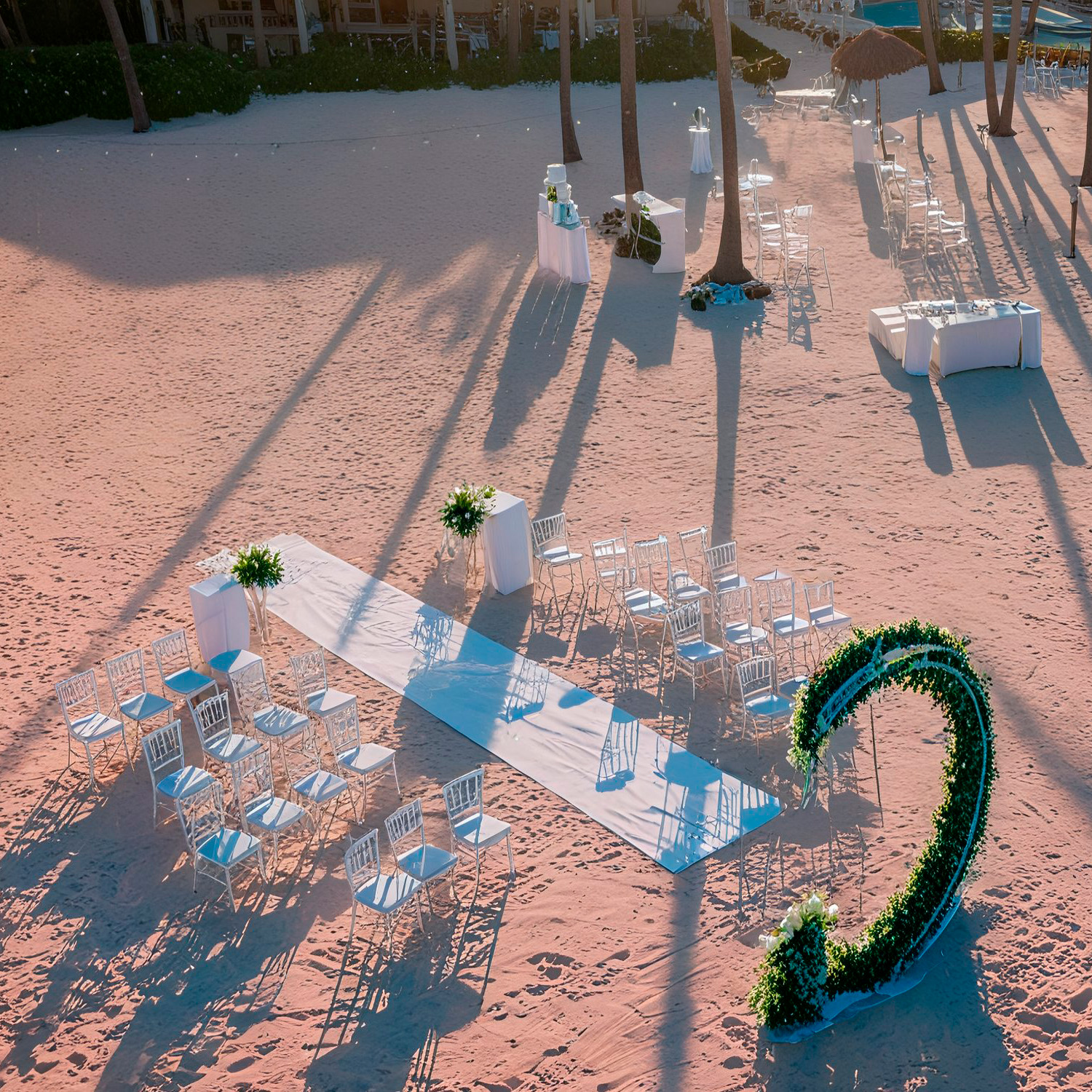An outdoor wedding setup on a sandy beach with white chairs arranged around an aisle, flanked by green plants and a large decorative number "2" adorned with greenery.